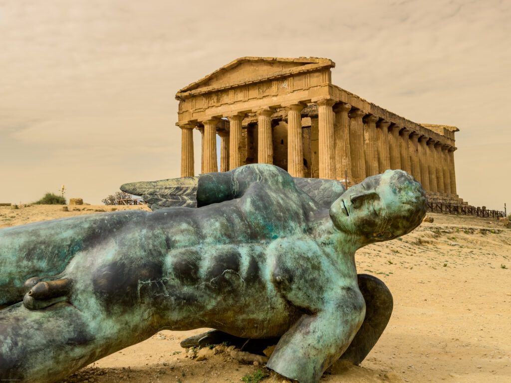 A fallen bronze statue in the foreground with an ancient temple in the background, set against a cloudy sky.